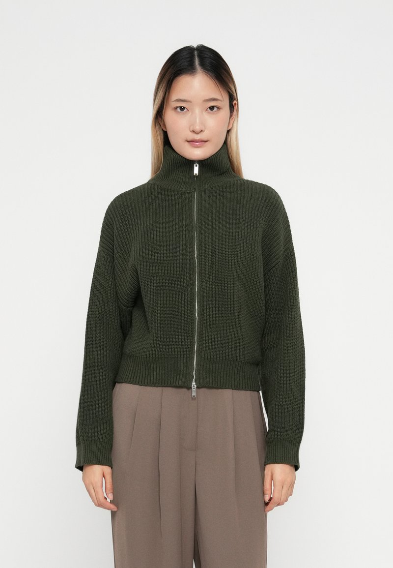 Maglione a coste di un verde scuro, con colletto alto e zip completa. Design cropped con maniche lunghe e tessuto testurizzato.
