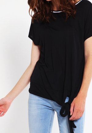 Haut noir ample avec des manches courtes, un col rond et une bordure blanche. Présente un détail de nouage froncé en bas.