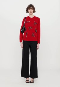 Red knitted sweater with black snake pattern details, paired with wide-leg black pants and a small black handbag.