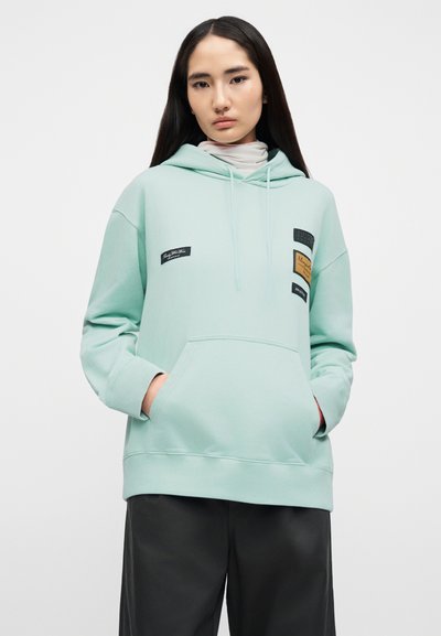 Φούτερ σε μέντα πράσινο με τσέπη καγκουρό, διακοσμημένο με διάφορα σημάδια, κουκούλα με κορδόνι και ριμπ μανσέτες. Φορεμένο με σκούρο παντελόνι.