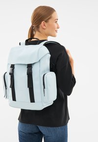 Hellblauer Rucksack mit schwarzen Riemen und Akzenten, ausgestattet mit einem oberen Deckel, zwei Seitentaschen und einem stabilen Griff. Glatte Textur.