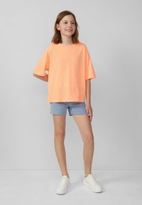 Neon-orange Oversize-T-Shirt mit kurzen Ärmeln, kombiniert mit hellblauen Denim-Shorts und weißen Sneakers, vor einem neutralen Hintergrund.
