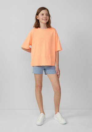 Neon oranje oversized T-shirt met korte mouwen, gedragen met lichtblauwe denim short en witte sneakers, tegen een neutrale achtergrond.