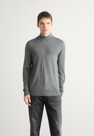 Grauer Rollkragenpullover aus glattem Strickstoff, mit gerippten Bündchen und Saum. Logo-Detail auf der Brust. Kombiniert mit dunklen Jeans.