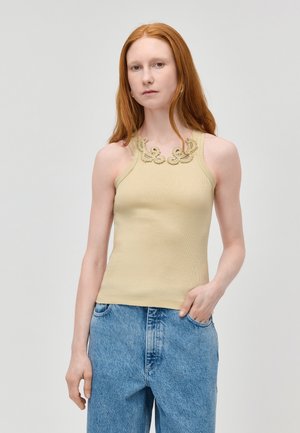 Femme aux cheveux roux portant un haut beige sans manches avec un détail en dentelle et un jean bleu ample, debout devant un fond clair uni.