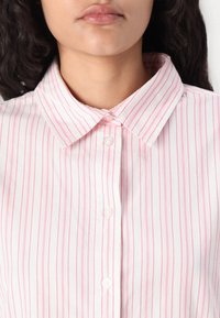 Camicia bianca con righe verticali sottili rosa, dotata di un colletto classico e bottoni bianchi lungo la patta anteriore. Tessuto liscio.