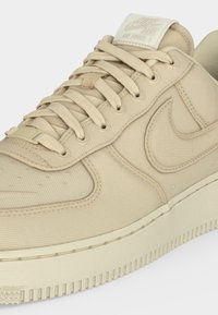Μπεζ αθλητικό παπούτσι Nike Air Force 1 με υφασμάτινη υφή, ραμμένο λογότυπο Nike, επενδεδυμένη γλώσσα και μπεζ κορδόνια που ταιριάζουν.