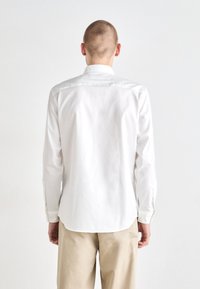 Chemise blanche à manches longues avec une texture lisse, dotée d'un col classique, d'un empiècement en dos et d'un ourlet arrondi. Portée avec un pantalon beige.