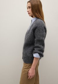 Cardigan grigio lavorato a maglia con una texture morbida, caratterizzato da maniche larghe e una vestibilità comoda, indossato sopra una camicia a righe blu e bianca.