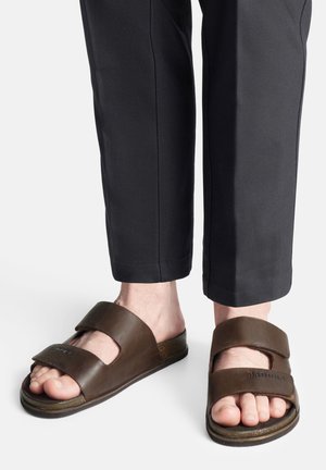 Brązowe skórzane klapki na płaskiej podeszwie z dwiema szerokimi paskami, posiadające regulowane zapięcia oraz teksturowaną powierzchnię, zestawione z dopasowanymi czarnymi spodniami.