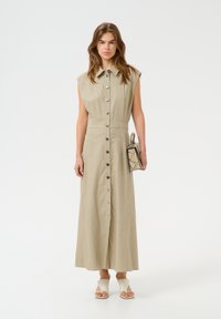 Abito camicia in lino khaki con maniche corte, chiusura frontale a bottoni e vita aderente. Indossato con sandali chiari e una piccola borsa con motivo a serpente.