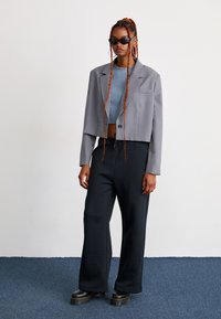 Giovane donna con lunghe trecce arancioni indossa un blazer grigio corto, un top blu corto, pantaloni larghi neri, scarpe chunky nere e occhiali da sole neri.