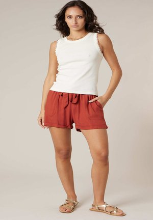 Woman wearing a white sleeveless top, rust-colored drawstring shorts, and beige strappy flat sandals, standing with one hand in pocket.