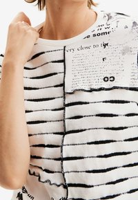 Dzianinowy top w paski w kolorze kremowym i czarnym, z różnorodnymi poziomymi paskami, teksturowaną tkaniną i nadrukowanymi detalami tekstowymi z jednej strony.