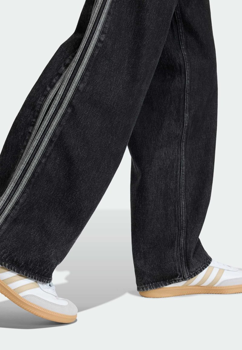 Schwarze Jeans-Hose mit weitem Bein und kontrastierenden grauen Seitenstreifen, kombiniert mit weißen Sportschuhen mit beigen Akzenten und drei grauen Streifen.