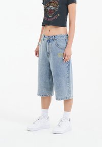 Culottes de mezclilla azul con un ajuste holgado, que presentan un detalle bordado en amarillo en la parte delantera, combinados con una camiseta gráfica negra recortada y zapatillas blancas.