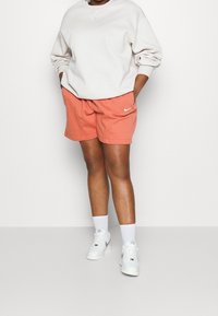 Ljusgrå Nike-tröja med rund hals, kombinerad med korallfärgade shorts med en sidologotyp. Vita strumpor och sneakers fulländar outfiten.