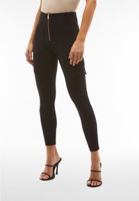 Leggings cargo neri a vita alta con zip frontale, dotati di tasche laterali e una vestibilità aderente. Materiale liscio e design alla caviglia.