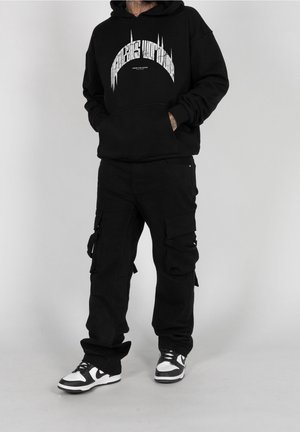 Persona con una sudadera gráfica negra, pantalones cargo negros y zapatillas en blanco y negro, de pie contra un fondo claro y liso.