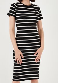 Donna che indossa un vestito nero aderente con strisce orizzontali bianche, maniche corte e scollatura rotonda, in piedi con le mani appoggiate sulle cosce.