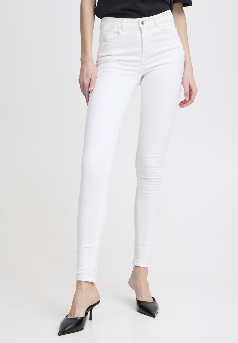 Jean skinny blanc à texture lisse, taille haute, avec une couture denim traditionnelle, associé à des escarpins noirs à bout pointu.