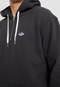 Musta vetoketjullinen huppari, joka on valmistettu puuvillasta ja jossa on valkoiset nyörit. Rinnassa on pieni sininen ja valkoinen logopatch. Pehmeä materiaali.