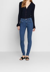 Top azul marino de manga larga con tejido acanalado, escote en V y cierre de botones, combinado con jeans ajustados azules y sandalias negras tipo slide.