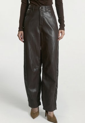 Persoon die donkerbruine leren wijde broek met hoge taille en voorzakken draagt, en bruine puntige pumps met hoge hakken.