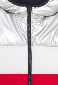 Puffjacka med en silver överdel, vit mitten och röd botten. Har en central dragkedja med svarta detaljer och en fodrad huva.