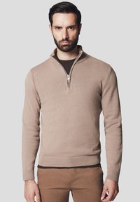 Beżowy sweter dzianinowy z kołnierzem na ćwierć zamka, prążkowanymi mankietami i dołem oraz gładką teksturą. Nosi się go na ciemno-brązowej koszuli.