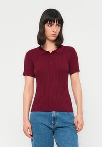 Donna con capelli scuri di media lunghezza che indossa una polo a maniche corte color bordeaux a coste e jeans in denim blu sullo sfondo neutro.