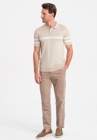 Beige stickad polo med ett vitt rand över bröstet, kombinerad med ljusbruna kordflädersbyxor och bruna slip-on skor.