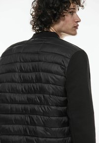 Svart jacka med en vadderad, quiltad kropp och ribbade ärmar. Slät textur med en figursydd design som visar en modern look.