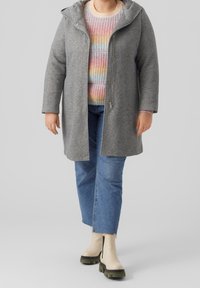 Grå uldfrakke med hætte, med lynlås foran, båret over en flerfarvet ribstrikket sweater og blå jeans med lige ben. Beige ankelstøvler.