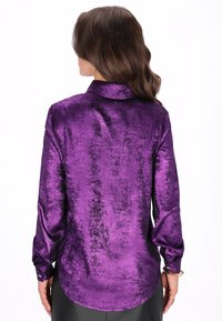 Lila hosszú ujjú ing, fényes textúrájú anyagból, galléros nyakkivágással és gombos mandzsettákkal. A hátsó részén lekerekített szegéllyel.