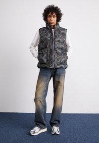 Padded camouflage vest s vysokým golierom, dve predné vrecká a zips. Skombinované so zodranými modrými džínmi a bielou športovou obuvou.