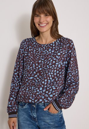 Femme souriante portant un haut marron à manches longues avec des taches abstraites bleues et un jean bleu, main dans la poche, sur un fond clair uni.