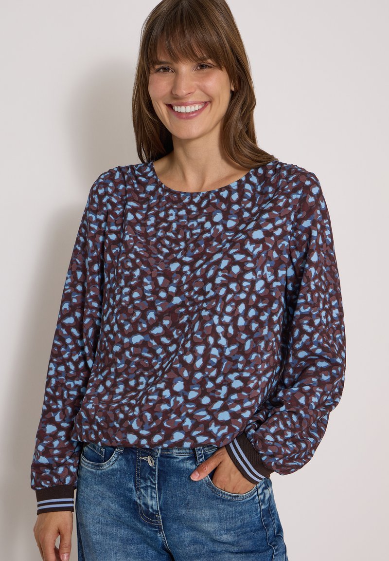 Femme souriante portant un haut marron à manches longues avec des taches abstraites bleues et un jean bleu, main dans la poche, sur un fond clair uni.