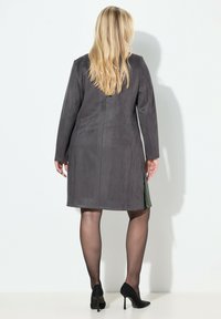 Abrigo de ante gris con mangas largas, corte recto y aberturas laterales. Zapatos negros de tacón alto contrastan con el conjunto. Textura suave con detalles mínimos.