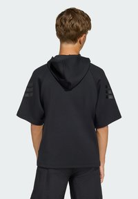 Ung person med kort brunt hår som bär en lös svart kortärmad hoodie med tre svarta ränder på varje ärm.