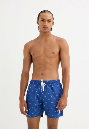 Joven con el cabello trenzado, vestido con pantalones cortos de baño azules con un patrón de delfines, de pie frente a un fondo blanco sencillo.