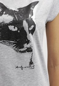Szary t-shirt z czarnym nadrukiem w postaci twarzy kota, z wyróżniającymi się oczami i wąsami, a obok znajduje się podpis wykonaną czarnym tuszem.