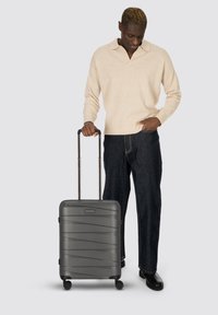 Uomo in maglione beige e jeans scuri in piedi con una valigia nera su ruote, che guarda in basso, una mano nella tasca, sfondo neutro.