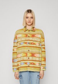 Vrouw met steil blond haar, gekleed in een kleurrijke geborduurde fleecejack met knopen en lichtblauwe cargobroeken, staand tegen een effen achtergrond.