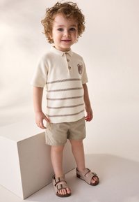 Beżowa dzianinowa koszulka polo z poziomymi brązowymi paskami, haftem misia i trzema guzikami. Do tego jasnobeżowe szorty i brązowe sandały.