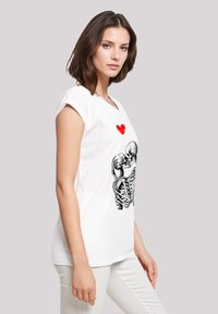 Weißes T-Shirt mit kurzen Ärmeln, das ein schwarzes Skelett-Design mit einem roten Herz darüber zeigt. Weicher Stoff mit lockerer Passform.