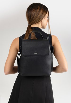 Donna con capelli castani raccolti in una coda di cavallo, che indossa una canotta nera e uno zaino in pelle nera con patta e piccolo logo dorato.