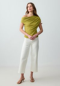Femme portant un top olive vert à épaules dénudées, un pantalon blanc à jambes larges et des sandales à talons bruns, se tenant devant un fond clair et uni.
