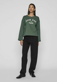 Sudadera verde con el texto blanco "VOUS ÊTES À PARIS", de mangas largas y corte relajado. Combinada con pantalones negros de pierna recta y zapatos negros.