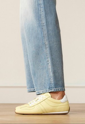 Persoon draagt lichtgele lage sneakers met witte veters en hieldetail, gecombineerd met lichtblauwe jeans die net boven de enkel zijn afgeknipt.